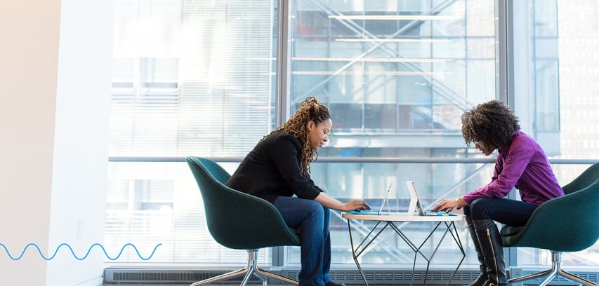 Two people use laptops seated at a small office lounge table