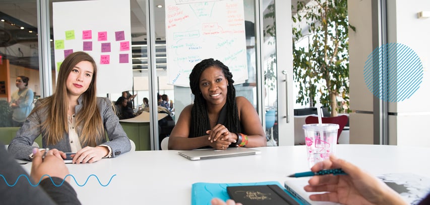 Two women sit on one side of a desk listening to someone speaking on the other side